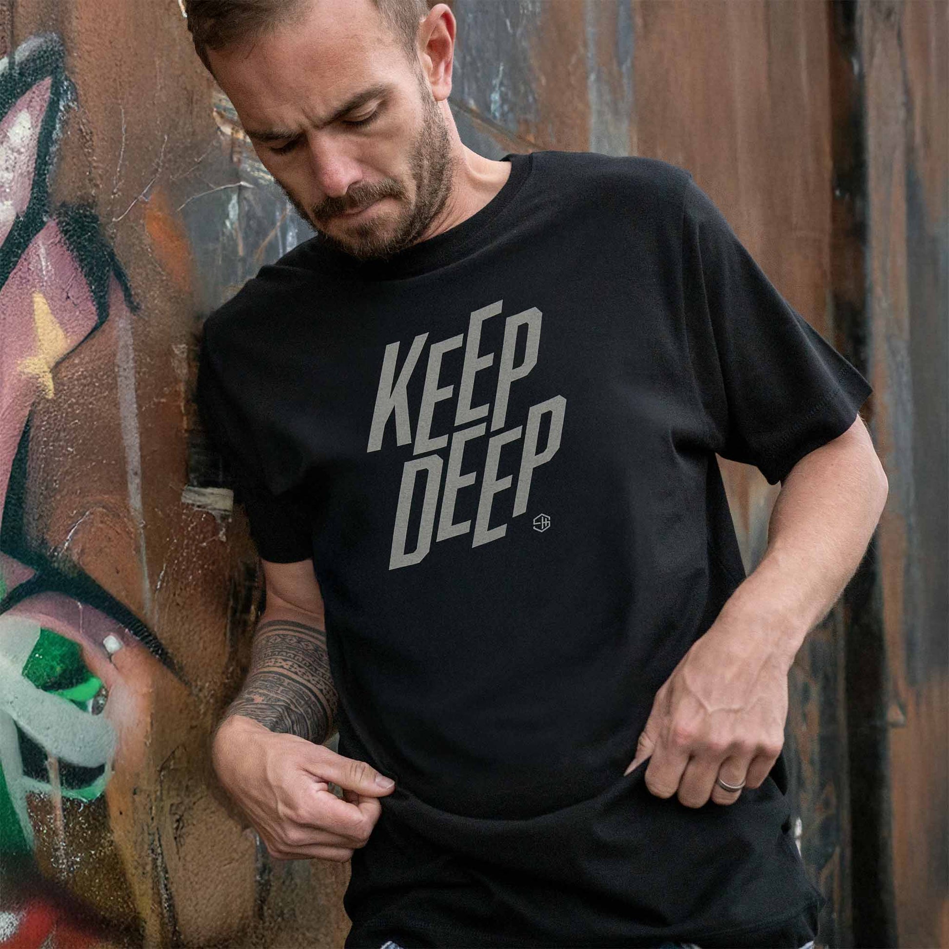 Man wearing a deep house black t-shirt with 'KEEP DEEP' text in front of a graffiti wall.