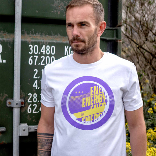 Man wearing a white house music t-shirt with a purple and yellow 90s graphic design, standing in front of a green container.
