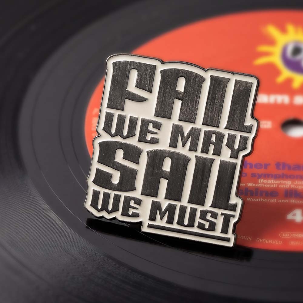 Metallic pin badge with 'Fail We May, Sail We Must' text on a vinyl record background