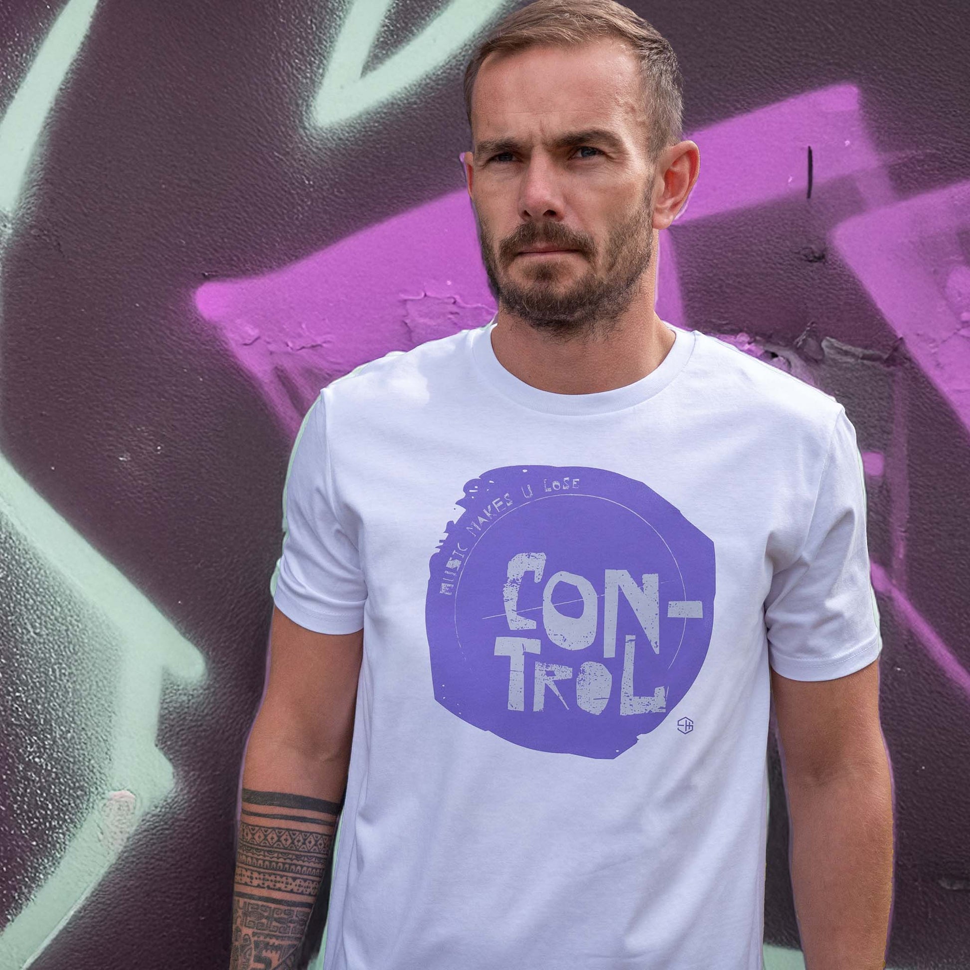 Man wearing a white house music t-shirt with a purple music makes you lose control graphic and text, standing in front of a colourful graffiti wall.