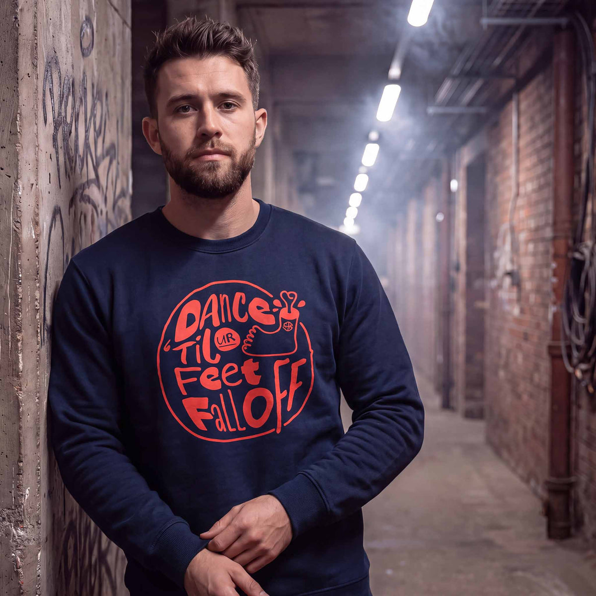Man wearing a navy dance music sweatshirt with red DANCING FEET screen print, dance ‘til ur feet fall off in a warehouse